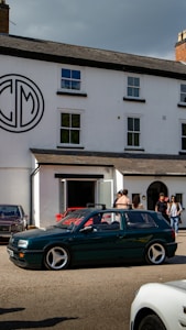 A group of people standing outside a white building with a large circular logo on the wall. Several cars, including a green station wagon with distinctive rims, are parked or driving by. The setting appears to be a gathering or meet-up on a clear day.
