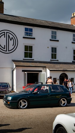 A group of people standing outside a white building with a large circular logo on the wall. Several cars, including a green station wagon with distinctive rims, are parked or driving by. The setting appears to be a gathering or meet-up on a clear day.