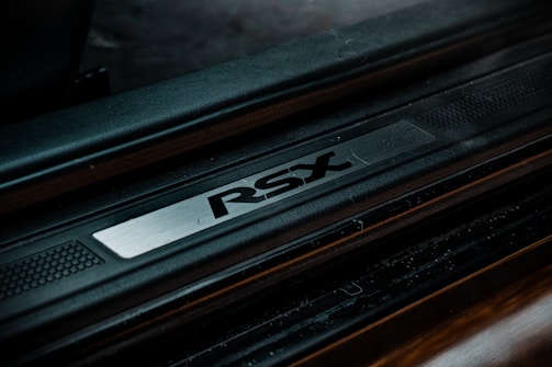 A close-up view of a car door sill plate with the letters 'RSX' prominently engraved on a metallic strip. The surrounding area is a textured black surface, suggesting a sleek automotive design.
