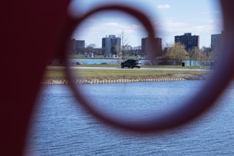 A lakeside scene is viewed through a circular metal structure, with a road running alongside the water. Several apartment buildings are visible in the background, along with some bare trees and bushes on a grassy area. A black SUV is seen driving on the road.