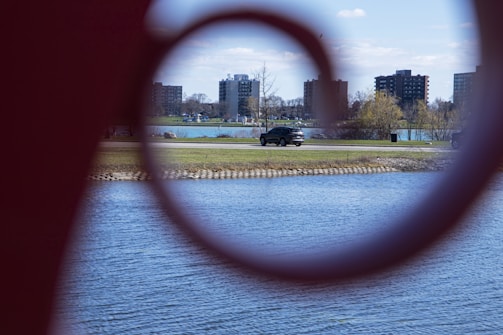 A lakeside scene is viewed through a circular metal structure, with a road running alongside the water. Several apartment buildings are visible in the background, along with some bare trees and bushes on a grassy area. A black SUV is seen driving on the road.