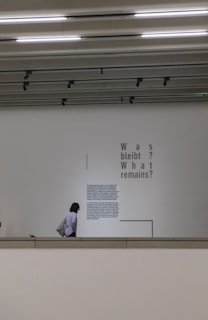 A person is walking past a wall with large text and a smaller block of writing. The larger text poses a question in two languages: 'Was bleibt? What remains?'. The setting appears to be indoors, possibly in a gallery or museum with overhead lighting and a minimalist design.