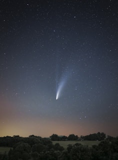 A night sky filled with stars and a bright comet with a long tail. Below the sky is a silhouette of trees and a landscape, creating a contrast with the illuminated comet.