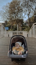 A baby dressed in a yellow sweater and white beanie lies comfortably inside a gray car seat placed on a wooden bridge. Trees with autumn foliage and a clear blue sky form the background, creating a serene atmosphere.