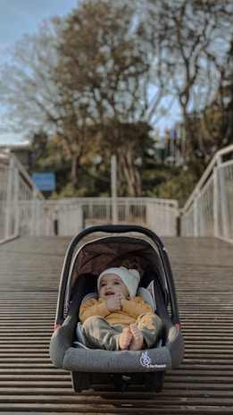 A baby dressed in a yellow sweater and white beanie lies comfortably inside a gray car seat placed on a wooden bridge. Trees with autumn foliage and a clear blue sky form the background, creating a serene atmosphere.