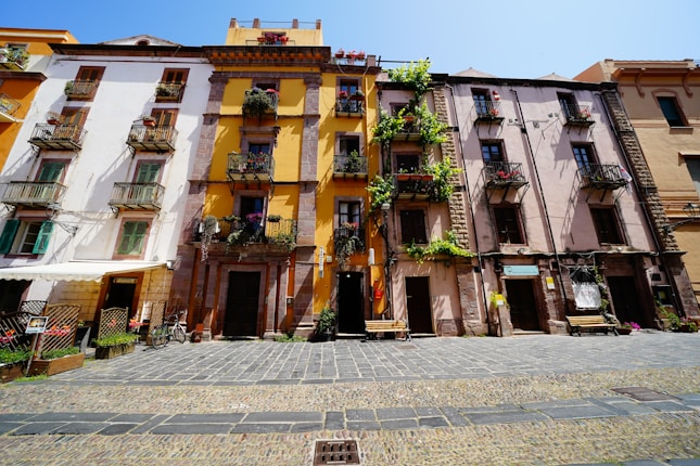 A charming street scene with colorful, historic European buildings featuring balconies adorned with flowers and greenery. The cobblestone pathway in front, along with benches and bicycles, adds to the quaint atmosphere.