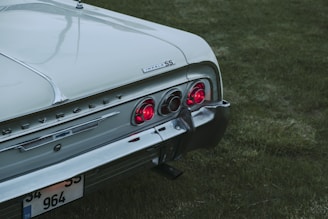 The rear section of a vintage Chevrolet Impala car is parked on grass. The vehicle is light-colored with shining chrome details, featuring distinctive red taillights and the Impala SS badge.