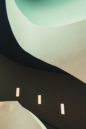 A modern architectural scene featuring curved, flowing lines and smooth surfaces. The design creates a sense of fluidity and movement, with a gradient of colors from light mint green transitioning to dark, muted tones. Rectangular lights are embedded in the walls, providing a soft, ambient glow.