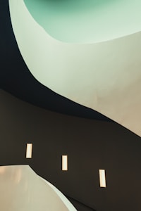 A modern architectural scene featuring curved, flowing lines and smooth surfaces. The design creates a sense of fluidity and movement, with a gradient of colors from light mint green transitioning to dark, muted tones. Rectangular lights are embedded in the walls, providing a soft, ambient glow.