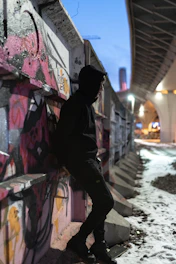 Model wearing a dark hoodie with urban artwork, standing under a graffiti-covered overpass at dusk.