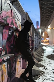 Urban young man wearing a black hoodie with neon green accents, standing against a graffiti wall at dusk.