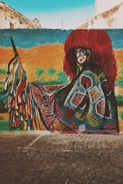 A colorful graffiti mural on a street wall depicts a person wearing a vibrant, patterned costume with a large, red headpiece. The background features a mix of bright colors like blue, yellow, and green, with a sky visible at the top. There are buildings in the distance, and the mural is located on a paved street.