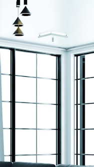 A modern interior setting featuring large, grid-style windows with black frames. The space is illuminated by geometric pendant lights hanging from the ceiling. A contemporary sofa is partially visible in the lower part of the image.