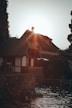 A picturesque lakeside scene featuring a large house with a steep roof and traditional architecture. The sun is setting behind the house, casting a warm glow and creating lens flares. Surrounding the house are lush trees, and the calm water in front reflects the light and the structure.