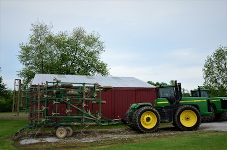 A large green tractor is positioned in front of a red barn with a metallic roof. The tractor is attached to a piece of farming equipment with multiple wheels. The background features lush green trees and a cloudy sky.