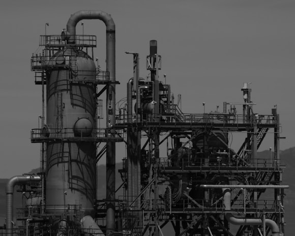 A large industrial structure featuring various pipes, tanks, and towers. The complex network of metal components creates a maze-like appearance, indicating a possible refinery or processing plant. The image is in black and white, enhancing the intricate details and shadows of the structure against a clear sky.