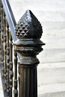 A close-up of a decorative cast iron railing post with a textured, pointed finial on top. The background features blurred stone steps, highlighting the intricate design and glossy finish of the post.