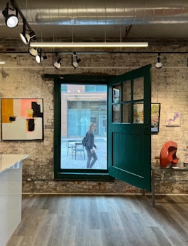 An interior of an art gallery with exposed brick walls and track lighting on the ceiling. There are several colorful abstract paintings on the walls and a red sculpture on a table to the right. The gallery has a large open window with a person visible outside walking.