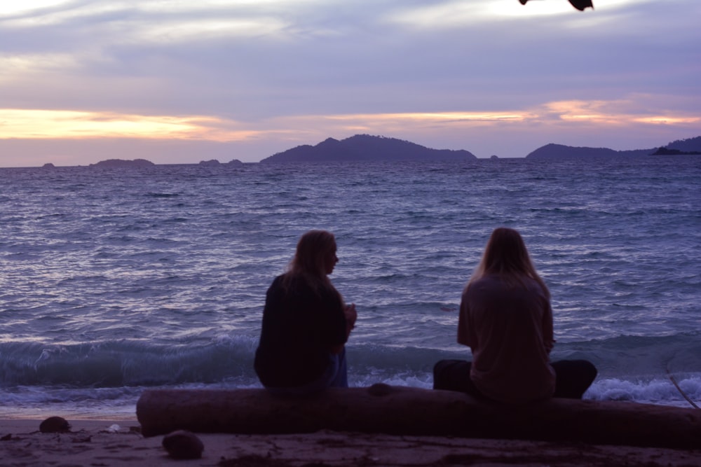 two people sitting on a log looking out at the ocean