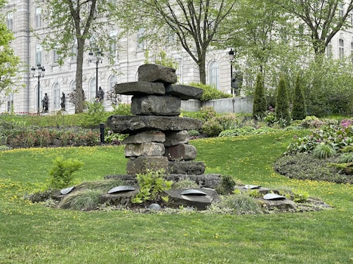 A stacked stone sculpture is surrounded by a well-maintained garden with lush greenery and flowering plants. In the background, a historic building with arched windows and statues is visible, flanked by mature trees and classic lampposts.