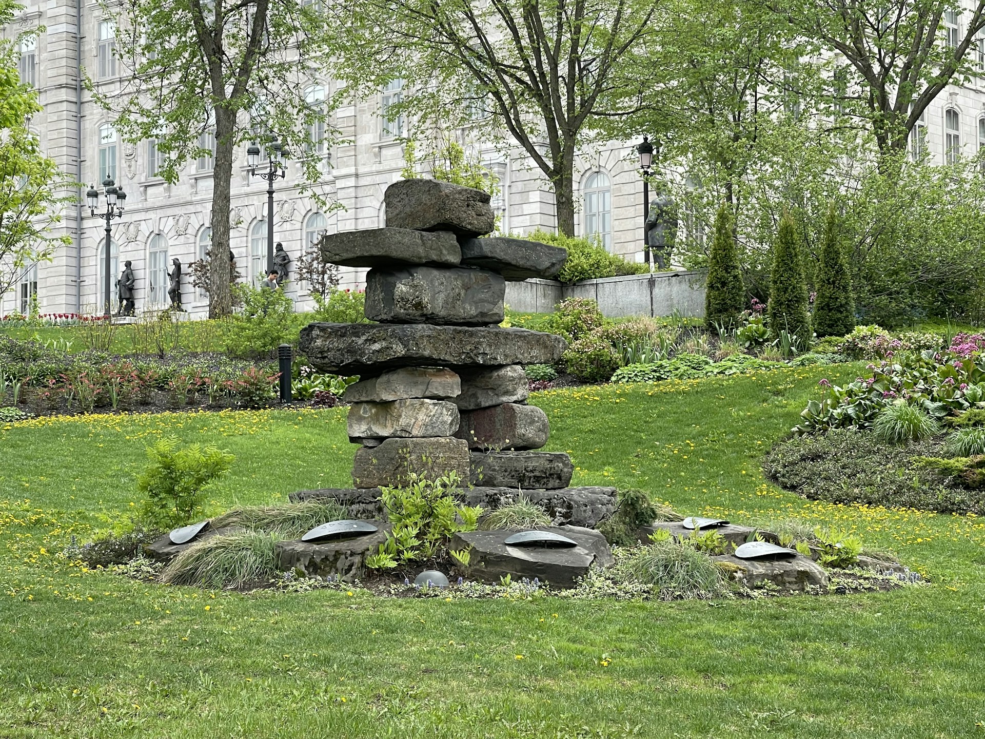 A stacked stone sculpture is surrounded by a well-maintained garden with lush greenery and flowering plants. In the background, a historic building with arched windows and statues is visible, flanked by mature trees and classic lampposts.