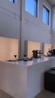 A minimalist coffee bar setup with clean, white surfaces and professional coffee-making equipment, including coffee drippers, placed on the counter. The space is illuminated by natural light from large windows, providing a calm and serene atmosphere.