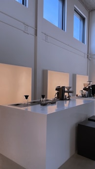 A minimalist coffee bar setup with clean, white surfaces and professional coffee-making equipment, including coffee drippers, placed on the counter. The space is illuminated by natural light from large windows, providing a calm and serene atmosphere.