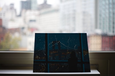 A piece of artwork resting on a window sill with a blurred cityscape visible through the glass. The painting depicts a nighttime city scene with abstract elements, including a bridge and lights. The background shows high-rise buildings in soft focus, suggesting an urban setting.