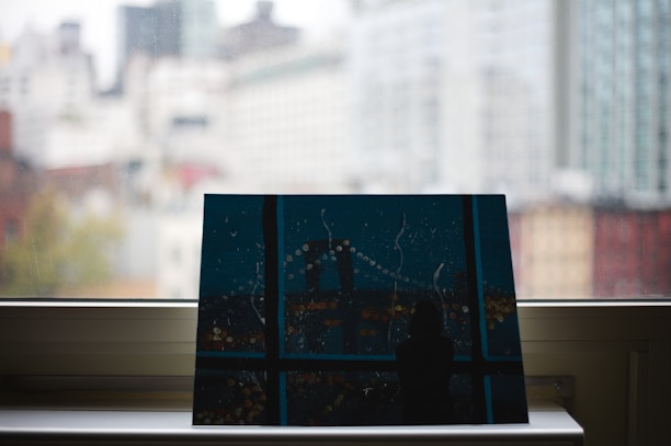 A piece of artwork resting on a window sill with a blurred cityscape visible through the glass. The painting depicts a nighttime city scene with abstract elements, including a bridge and lights. The background shows high-rise buildings in soft focus, suggesting an urban setting.