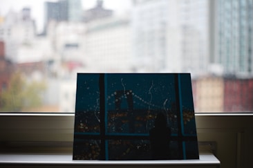 A piece of artwork resting on a window sill with a blurred cityscape visible through the glass. The painting depicts a nighttime city scene with abstract elements, including a bridge and lights. The background shows high-rise buildings in soft focus, suggesting an urban setting.