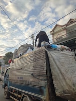 A truck loaded with various items, including bags and debris, is parked on the street. A person is standing on top of the truck, seemingly arranging or sorting through the load. The scene takes place in an urban setting with residential buildings visible in the background. Overhead, power lines crisscross the sky, which is partly cloudy.