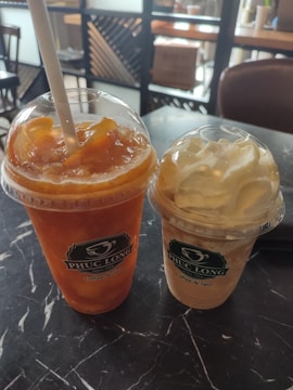Two plastic cups with dome lids containing beverages are placed on a dark marble table. The left cup holds an iced tea with slices of orange and a straw, while the right cup contains a creamy drink topped with whipped cream.
