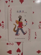 A playing card is prominently displayed with an illustration of a joker character. The character wears a crown, a red outfit, and has a dragon tail. It is set against a backdrop of scattered playing cards featuring red suits such as hearts and diamonds.