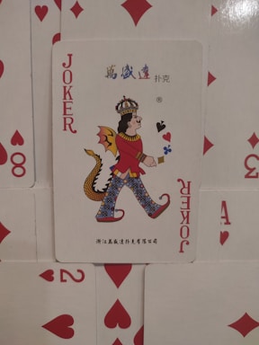 A playing card is prominently displayed with an illustration of a joker character. The character wears a crown, a red outfit, and has a dragon tail. It is set against a backdrop of scattered playing cards featuring red suits such as hearts and diamonds.