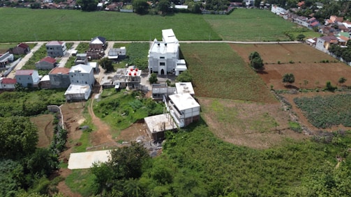 Aerial view of a rural area featuring a large white building, possibly a church, surrounded by smaller residential structures. The landscape consists of green fields with patches of brown soil, indicating agricultural land. Trees and vegetation are scattered throughout, and a dirt road runs through the area. In the background, more houses and farmland can be seen.