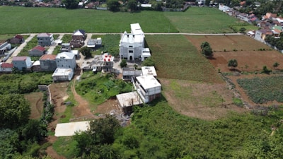 Aerial view of a rural area featuring a large white building, possibly a church, surrounded by smaller residential structures. The landscape consists of green fields with patches of brown soil, indicating agricultural land. Trees and vegetation are scattered throughout, and a dirt road runs through the area. In the background, more houses and farmland can be seen.