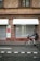 A brick building with a closed shop named Johann Schuhe, featuring two awnings above display windows covered by metal shutters. A person rides a bicycle on the road in front of the shop.