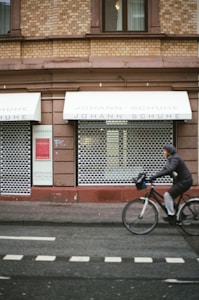 A brick building with a closed shop named Johann Schuhe, featuring two awnings above display windows covered by metal shutters. A person rides a bicycle on the road in front of the shop.