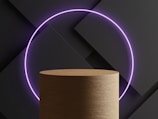 A cylindrical wooden platform is positioned in front of a modern, geometric background with overlapping dark panels. A glowing purple neon circle is centrally placed in the composition, creating a futuristic and minimalist aesthetic.