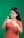 A person with long, dark hair is smiling while giving double thumbs up. They are wearing an orange patterned top and are positioned against a plain green background.