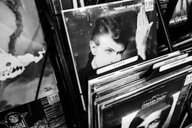 A black-and-white photograph of several vinyl records displayed in a store. Prominently featured is a record with an image of a man striking a dramatic pose, surrounded by other albums with visible price tags and artistic covers. The environment suggests a vintage or retro aesthetic.