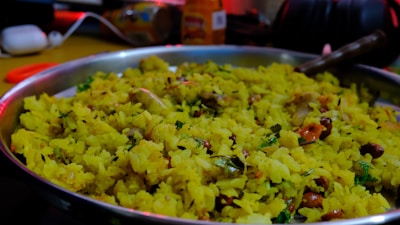 A close-up of a silver plate filled with yellow poha, a flattened rice dish. The poha is mixed with spices, garnished with coriander leaves, and has visible pieces of peanuts and curry leaves. The background is slightly blurred with some kitchen items.