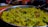 A close-up of a silver plate filled with yellow poha, a flattened rice dish. The poha is mixed with spices, garnished with coriander leaves, and has visible pieces of peanuts and curry leaves. The background is slightly blurred with some kitchen items.
