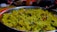 A close-up of a silver plate filled with yellow poha, a flattened rice dish. The poha is mixed with spices, garnished with coriander leaves, and has visible pieces of peanuts and curry leaves. The background is slightly blurred with some kitchen items.