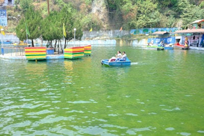 A pond with people enjoying paddle boats on calm green water. There are several small floating platforms with colorful striped barriers and trees. The background features a lush hillside and a mural on a wall. A dock with additional paddle boats is present to the side.