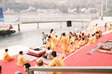 A group of people dressed in orange and yellow traditional attire are gathered near a riverbank. They are seated on red and patterned mats on steps leading down to the water. Some individuals appear to be engaged in religious or spiritual practices. A wooden platform extends into the river, and further in the background, hills and buildings are visible across the water.