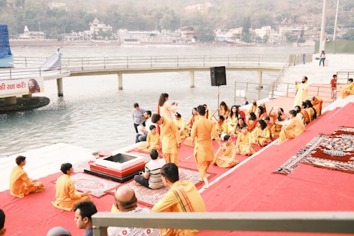 A group of people dressed in orange and yellow traditional attire are gathered near a riverbank. They are seated on red and patterned mats on steps leading down to the water. Some individuals appear to be engaged in religious or spiritual practices. A wooden platform extends into the river, and further in the background, hills and buildings are visible across the water.