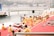 A group of people dressed in orange and yellow traditional attire are gathered near a riverbank. They are seated on red and patterned mats on steps leading down to the water. Some individuals appear to be engaged in religious or spiritual practices. A wooden platform extends into the river, and further in the background, hills and buildings are visible across the water.