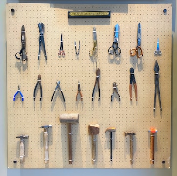 A pegboard is neatly organized with various tools, including scissors, pliers, hammers, and other hand tools. Each tool is secured on the board with a white strap. At the top, a label reads 'TOOLS OF THE TRADE'. The arrangement is symmetrical and orderly, with tools of different shapes and sizes.