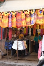 A vibrant street market stall featuring an array of colorful dresses hanging prominently at the front. The dresses are primarily in shades of orange, yellow, red, and blue, with intricate patterns. Beneath the dresses, there are racks of shorts and more clothing items. Two people are visible inside the stall, adding a lively human element to the scene.